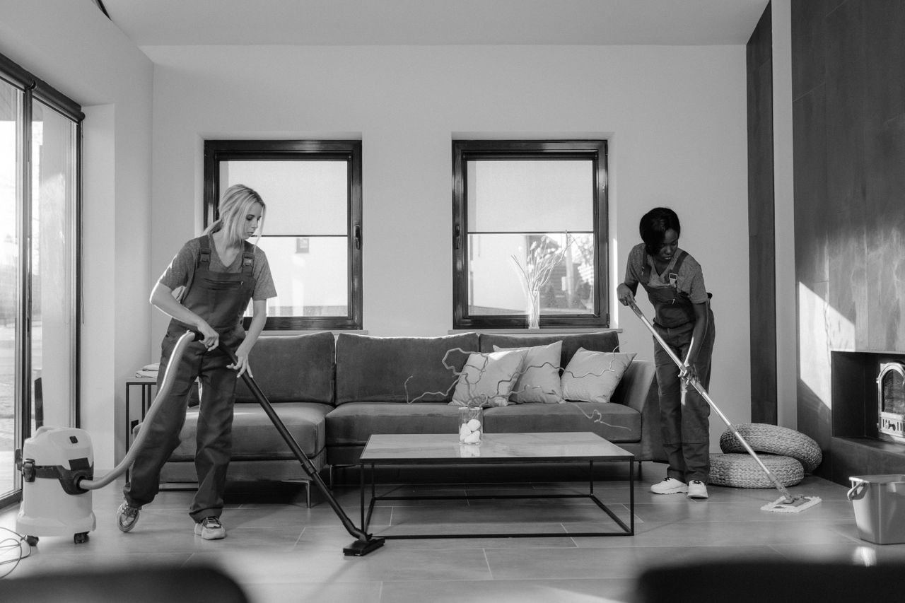 team-02 Two women in work clothes cleaning a modern living room using a vacuum and mop.